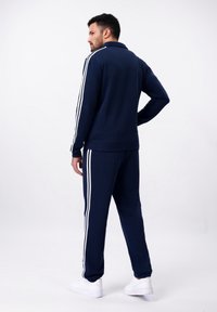 Survêtement marine avec une veste zippée et un pantalon assorti. Présente trois rayures blanches le long des manches et des pantalons. Texture douce.