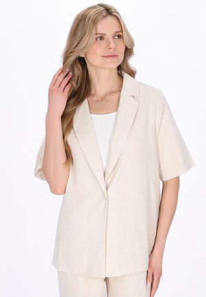 Hosszú szőke hajú nő, bézs rövid ujjú blézert és hozzá illő nadrágot viselve, a haját érintve, oldalra nézve.