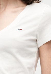 Κοντινό πλάνο ενός ατόμου που φοράει λευκό μπλουζάκι με V λαιμόκοψη και κοντά μανίκια, με ένα μικρό κόκκινο, λευκό και μπλε ορθογώνιο λογότυπο στο στήθος.