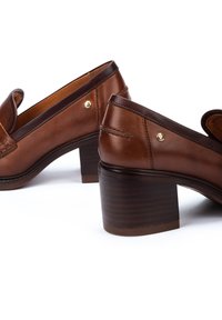 Mocasines de cuero marrón con tacón, punta redondeada, costuras decorativas y un tacón de bloque bajo con textura de madera. Detalle en dorado en el lateral.