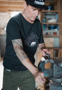 Camiseta negra con un diseño de oso geométrico, pantalones verde oliva, tatuaje de brazo bien definido, trabajando con un tornillo metálico en un taller.