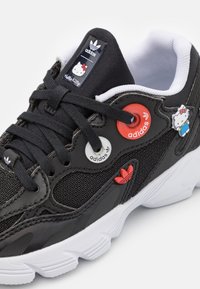 Zapatilla deportiva negra texturizada con suela blanca, que presenta adornos de Hello Kitty y acentos coloridos del logo de Adidas en la parte superior.