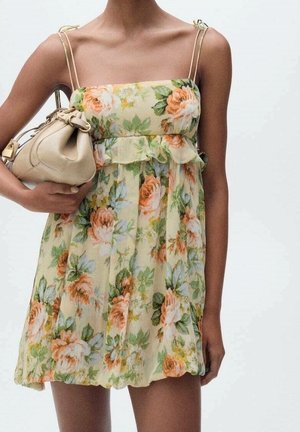 Femme portant une robe jaune à fleurs sans manches, tenant un sac à main beige sous le bras sur un fond uni.