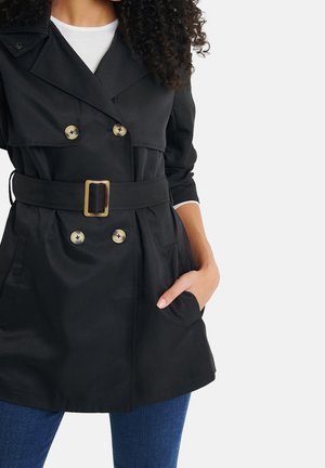 Vrouw die een zwarte double-breasted trenchcoat draagt met gouden knopen en riem, wit shirt eronder en blauwe spijkerbroek, hand in de zak.