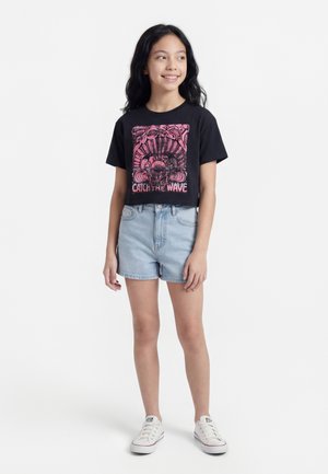 Κορίτσι που στέκεται φορώντας μαύρο crop top με ροζ γράφημα "Catch the Wave", ανοιχτόχρωμο τζιν σορτς και λευκά αθλητικά παπούτσια, χαμογελώντας και κοιτάζοντας δεξιά.