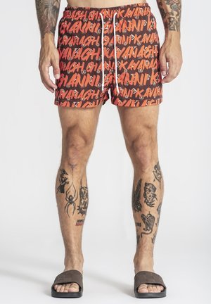Homme portant un short rouge et noir à motifs avec le texte « Gianni Kavanagh », des tatouages sur les jambes et les bras, et des sandales marron sur fond blanc.
