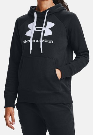 Osoba ubrana w czarną bluzę z kapturem Under Armour z białym logo i sznurkami, ręka w przedniej kieszeni, na jednolitym białym tle.