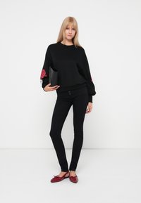 Sudadera negra con estampado de rosas rojas en las mangas, combinada con jeans skinny negros y zapatos planos rojos. La modelo sostiene un clutch negro.