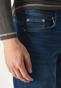 Jeans de mezclilla azul con costuras claras, que presentan un pequeño bolsillo con botones de metal. La mano está colocada en el costado de los jeans.