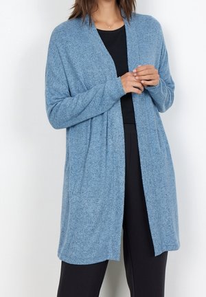 Frau trägt eine weiche, hellblaue Cardigan mit offenem Vorderteil über einem schwarzen Oberteil und schwarzen Hosen, steht vor einem einfachen weißen Hintergrund.