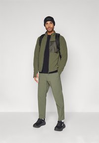 Giacca in pile verde oliva con una tasca scura, t-shirt nera, pantaloni verde oliva, berretto nero e scarpe da ginnastica nere. Zaino incluso.