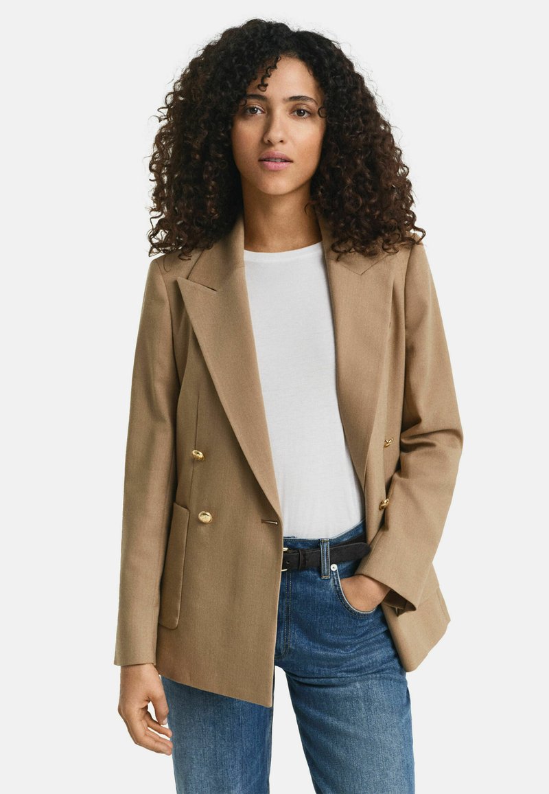 Blazer beige doppiopetto con bottoni dorati, rever a contrasto e due tasche frontali, indossato sopra una camicia bianca e jeans blu.