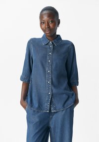 Mujer de piel oscura vestida con una camisa de mezclilla azul y pantalones de mezclilla a juego, de pie con las manos en los bolsillos contra un fondo blanco.