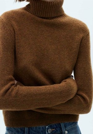 Pull à col roulé marron côtelé porté avec un jean en denim bleu, les bras croisés, sur un fond clair uniforme.