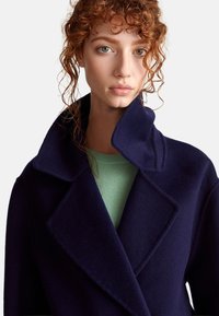 Cappotto di lana blu navy con un ampio bavero, indossato sopra un top verde chiaro. Texture morbida con vestibilità su misura e linee pulite, che enfatizzano la struttura.