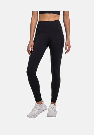 Femme portant un legging noir taille haute avec des poches latérales et des chaussures de sport blanches, debout devant un fond uni.