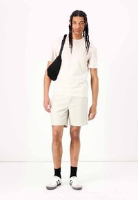 Beige short-sleeve t-shirt, light gray shorts, black crossbody bag, white sneakers with black accents, worn with black socks, neutral background.