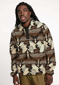 Pull en polaire avec un col montant, présentant un motif abstrait marron, crème et gris. Comprend une patte de boutonnage noire et une surface texturée.