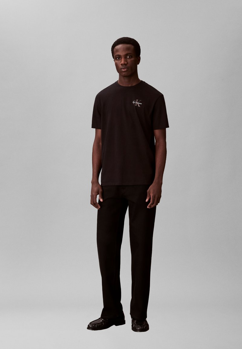 Άνδρας που φοράει μαύρο t-shirt Calvin Klein, μαύρο παντελόνι και μαύρα παπούτσια μπροστά σε απλό γκρι φόντο.