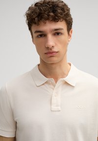 Crème poloshirt met een gestructureerde kraag, twee-knopen bies en een textuurstof. Heeft een subtiele, ingedrukte logo op de borst.