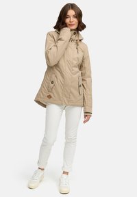 Beige jakke med hætte, lynlås foran, to sidelommer med knaplukninger, tekstureret stof, båret over hvide bukser og sneakers.