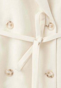 Gros plan sur un tissu beige avec quatre boutons ronds et une ceinture en tissu nouée passant par une fente sur un côté.
