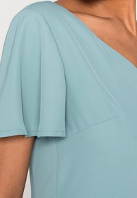 Jasnoniebieska sukienka z krótkimi falbaniastego rękawami, z dekoltem w kształcie litery V i gładką teksturą materiału. Na korpusie widoczne są szwy.