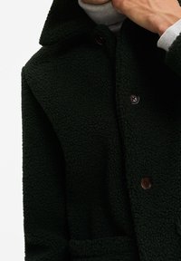 Tmavě zelený fleecový kabát s límečkem, dvěma knoflíky a strukturovaným povrchem. Obsahuje boční kapsu s jemným povrchem.