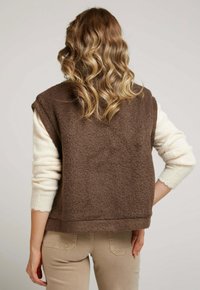 Gilet marron texturé avec des manches crème, présentant une forme boxy et des finitions côtelées. Superposé sur un pantalon beige, mettant en avant un style décontracté.