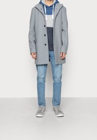 Grijze wollen jas met een capuchon, met een rechte pasvorm, voorknopen en zijzakken, gelaagd met een blauw-grijze gestreepte hoodie, gedragen met lichte jeans.