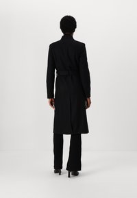 Cappotto lungo nero dalla vestibilità sartoriale, maniche lunghe, cintura in vita e spacco posteriore. Abbinato a pantaloni neri a costine e tacchi.