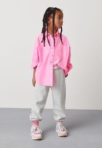 Bright pink oversized button-up shirt, gray sweatpants, and multicolored sneakers. Braided hair and a large front pocket on the shirt.