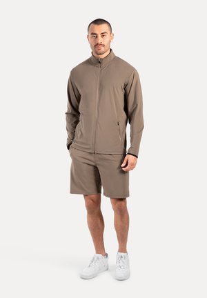 Ensemble de vêtements de sport pour hommes dans un marron terreux, comprenant une veste zippée et un short, tous deux en tissu lisse et extensible. Des baskets blanches complètent le look.