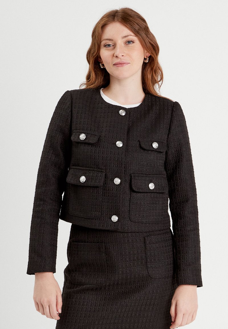 Zwarte cropped jas van textuurstof met zilveren knoopsluitingen, voorzien van twee borstzakken en een bijpassende rok, minimalistisch ontwerp.
