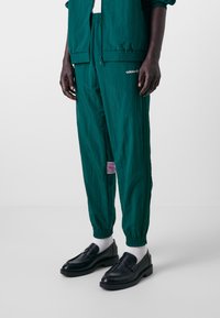 Calças de atletismo em azul-turquesa com um cós elástico e um bolso lateral, com um detalhe em roxo e uma marca discreta, combinadas com mocassins pretos e meias brancas.