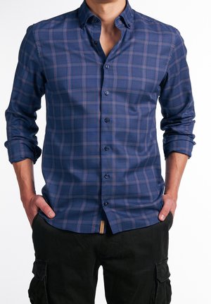 Mężczyzna ubrany w niebieską, długo rękawną koszulę w kratę z podwiniętymi rękawami, ręce w kieszeniach, połączoną z czarnymi spodniami typu cargo.