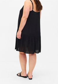 Žena ve bezramenných černých šatech s volánkovým lemem a černými sandály na klínu se špičatými hroty, stojící proti jednoduchému bílému pozadí.