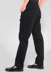 Zwarte broek van gladde stof, met een rechte pijp, twee zijzakken en een eenvoudig ontwerp van de tailleband. G draagt met zwarte schoenen.