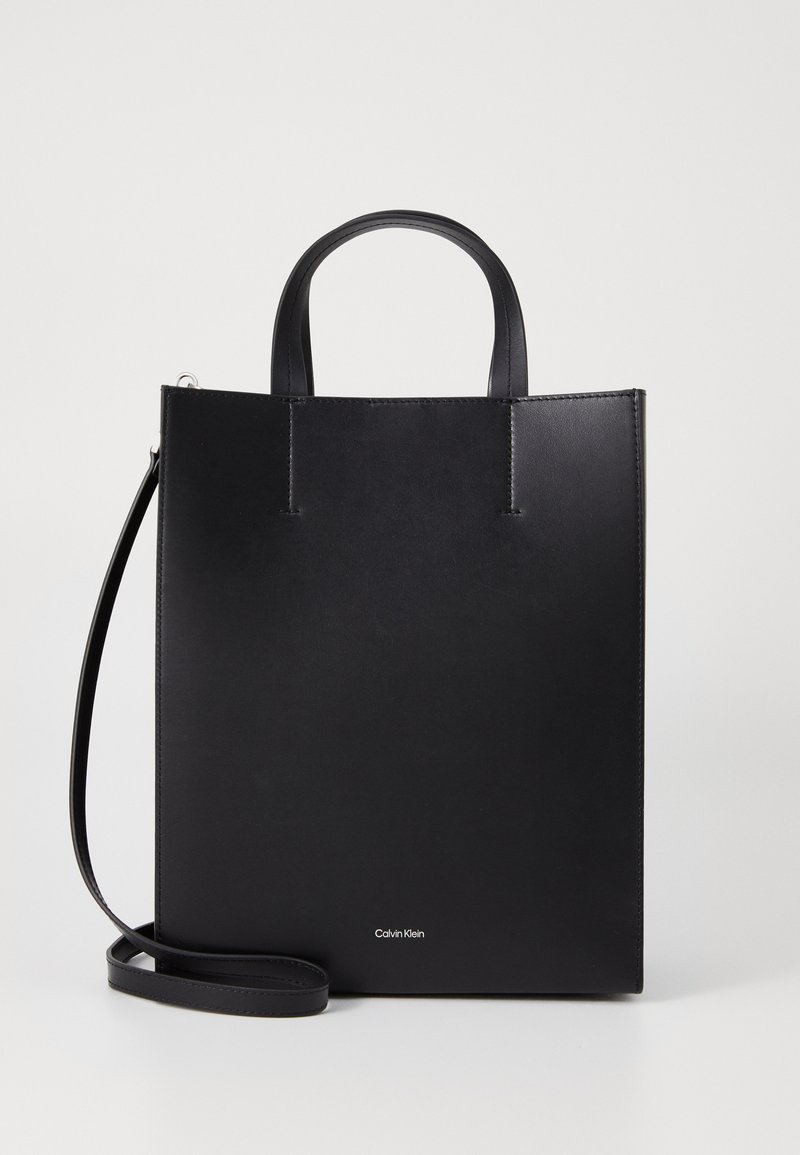 Geantă tote din piele neagră, cu două mânere superioare și o curea de umăr detașabilă. Prezintă un design minimal și un logo mic în partea de jos.