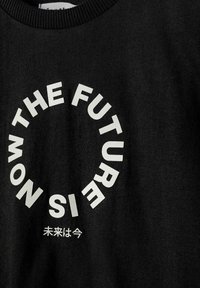 Tissu noir avec du texte blanc formant un cercle disant « MAINTENANT LE FUTUR EST » et de petits caractères japonais en dessous signifiant « Le futur, c'est maintenant. »