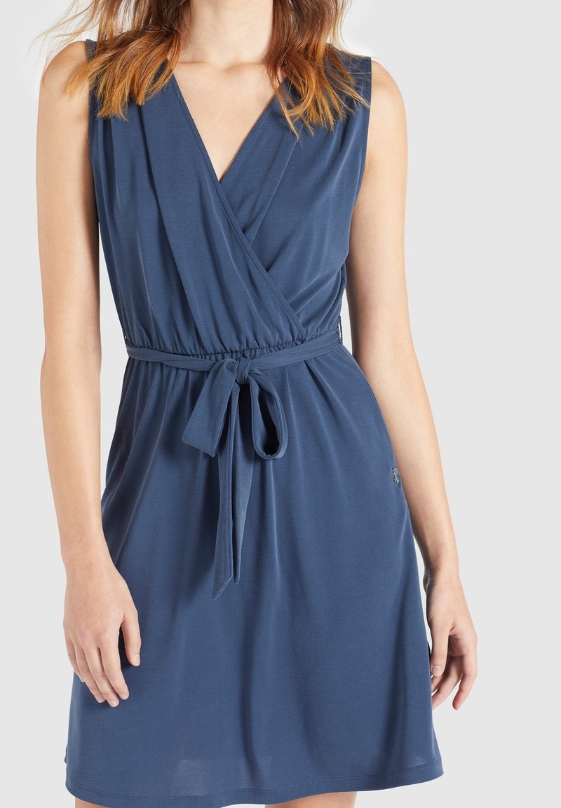 Femme portant une robe portefeuille bleue sans manches avec un col en V et une ceinture nouée à la taille sur un fond clair uni.