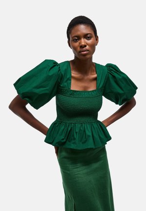 Femme aux cheveux courts portant un chemisier vert foncé à manches bouffantes et une jupe assortie, debout les mains sur les hanches devant un fond uni.