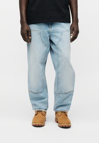 Άνδρας με ανοιχτό μπλε φαρδιά τζιν με μεγάλες μπροστινές τσέπες και ταμπα μποτάκια εργασίας με κορδόνια, στέκεται μπροστά σε απλό φόντο.