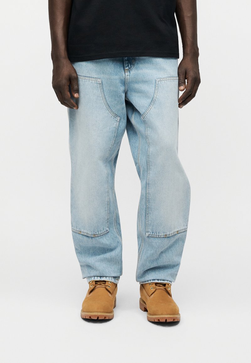 Homme portant un jean ample bleu clair avec de grandes poches avant et des bottes de travail à lacets couleur camel, debout devant un fond uni.