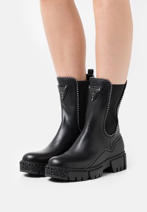 Black ankle boots made of leather with a chunky rubber sole. Features triangular metal logo, studded accents, and elastic side panels.