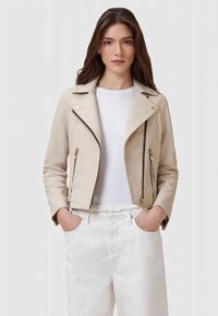 Mulher com cabelo castanho longo veste uma jaqueta bege claro com fecho éclair sobre uma camisa branca e calças brancas, com as mãos nos bolsos, contra um fundo simples.