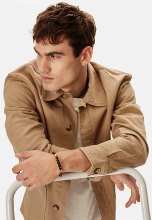 Giovane uomo con giacca beige appoggiato a una struttura metallica bianca, indossa un braccialetto nero con perline, guarda verso sinistra con sfondo semplice.