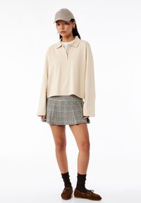 Beige langærmet polo shirt med krave, parret med en ternet plisseret nederdel. Tilbehør inkluderer en ternet cap og brune loafers.