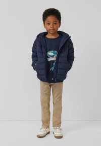 Marineblaue Pufferjacke mit Kapuze, beige Hose und weiße Sneakers. Das T-Shirt zeigt ein buntes Dinosaurier-Motiv.