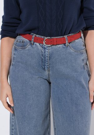 Persona che indossa un maglione blu lavorato a maglia e jeans azzurri a vita alta con una cintura rossa in suede con fibbia argentata, mani rilassate ai lati.